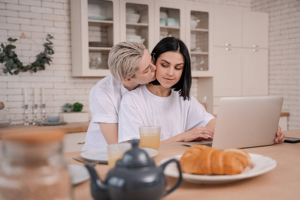 young woman kissing girlfriend while using laptop in kitchen  - Φωτογραφία, εικόνα