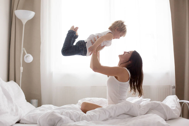 Beautiful blond child and his mother, playing at home in the morning in bed, smiling, laughing and having fun together - Фото, зображення