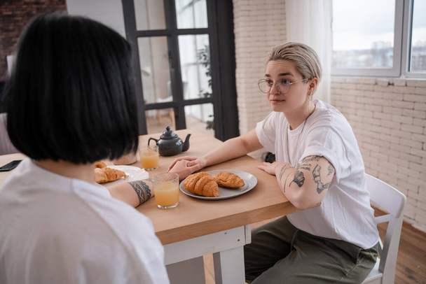 tattooed lesbian couple holding hands and looking at each other in kitchen  - Valokuva, kuva