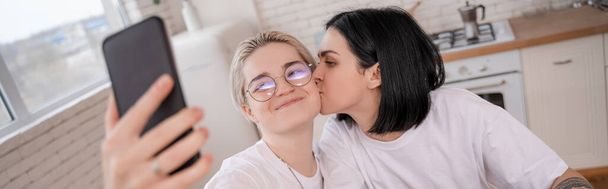morena mulher beijando bochecha de namorada tomando selfie na cozinha, banner - Foto, Imagem