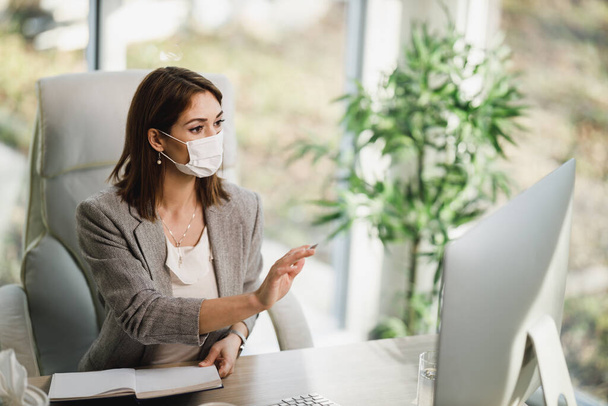 A young business woman with surgical mask sitting alone in her office and working on computer. - Foto, Imagen