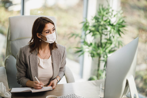Привлекательная задумчивая деловая женщина в хирургической маске сидит одна в офисе и работает за компьютером. - Фото, изображение