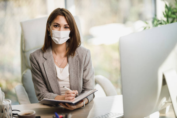 Eine erfolgreiche Geschäftsfrau mit OP-Maske sitzt allein in ihrem Büro und blickt in die Kamera. - Foto, Bild