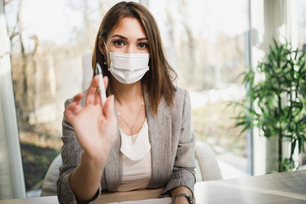 Een aantrekkelijke jonge vrouw met chirurgisch masker zittend in haar kantoor en pratend met haar cliënt. Kijkend naar camera. - Foto, afbeelding