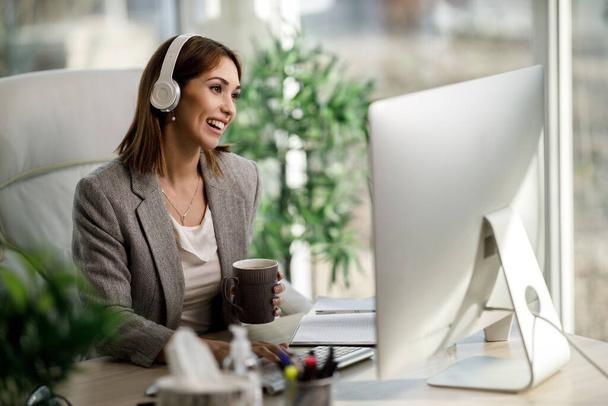 Una mujer de negocios sonriente disfrutando de una taza de café mientras trabaja en la computadora en la oficina en casa. - Foto, imagen