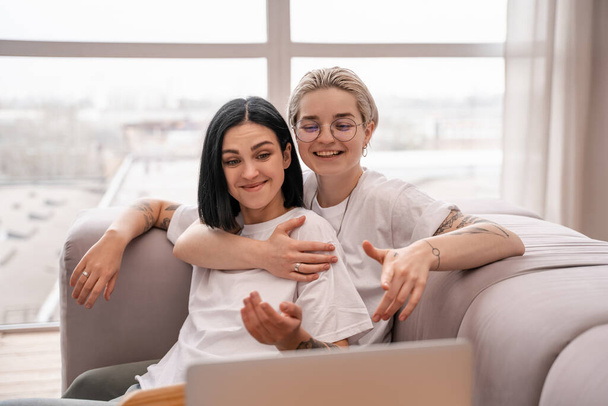 jeune couple lesbien souriant tout en refroidissant sur le canapé près d'un ordinateur portable  - Photo, image
