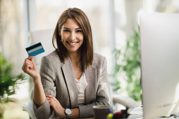 Una mujer de negocios sonriente sosteniendo una tarjeta de crédito y mirando a la cámara mientras trabaja en su computadora en la oficina. - Foto, imagen