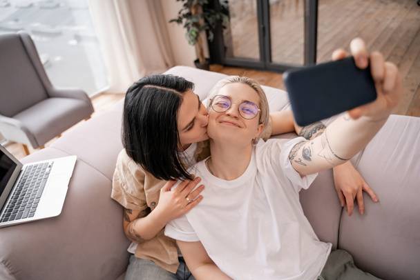 woman kissing cheek of happy girlfriend in glasses taking selfie in living room  - Φωτογραφία, εικόνα