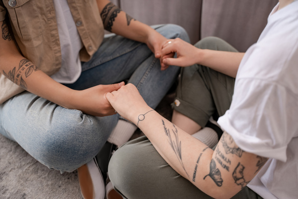 ostříhaný pohled na tetované lesbické páry sedící a držící se za ruce v obývacím pokoji  - Fotografie, Obrázek