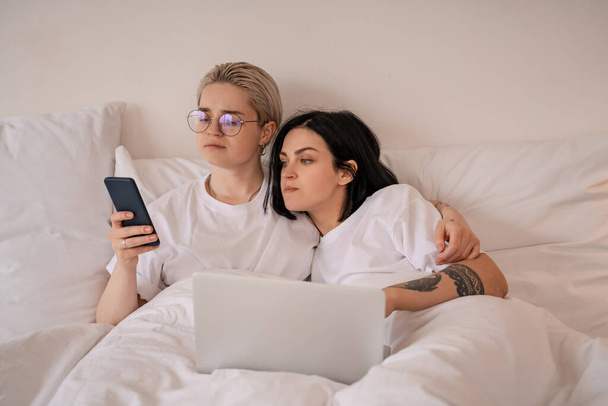 młoda lesbijska para leżąca w łóżku i patrząca na smartfona w pobliżu laptopa  - Zdjęcie, obraz
