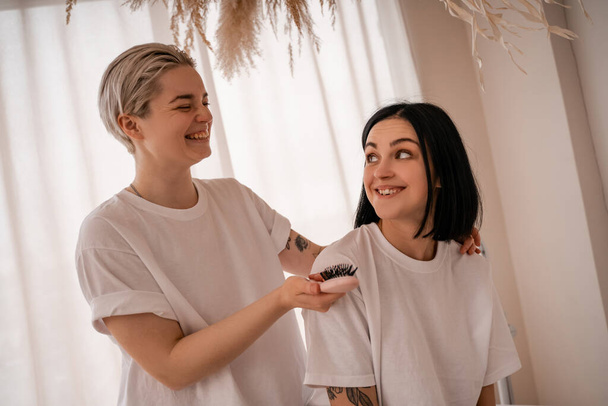 Neşeli genç bir kadın yatak odasında esmer kız arkadaşının saçlarını tarıyor.  - Fotoğraf, Görsel