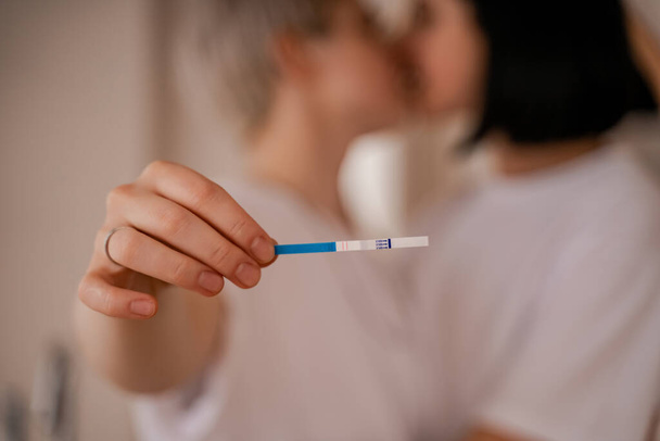 jong lesbisch vrouw holding zwangerschap test en zoenen vriendin op wazig achtergrond  - Foto, afbeelding