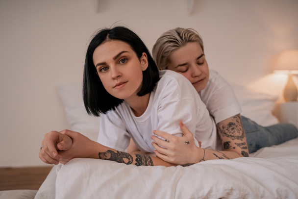 young lesbian woman hugging tattooed girlfriend and lying on bed - Photo, Image