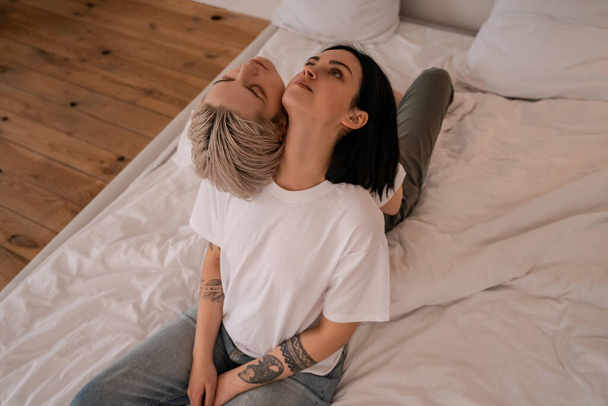 high angle view of tattooed lesbian couple sitting back to back on bed - Zdjęcie, obraz