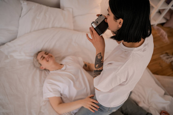 femme tatouée prenant une photo de petite amie floue allongée sur le lit et souriant sur la caméra rétro  - Photo, image