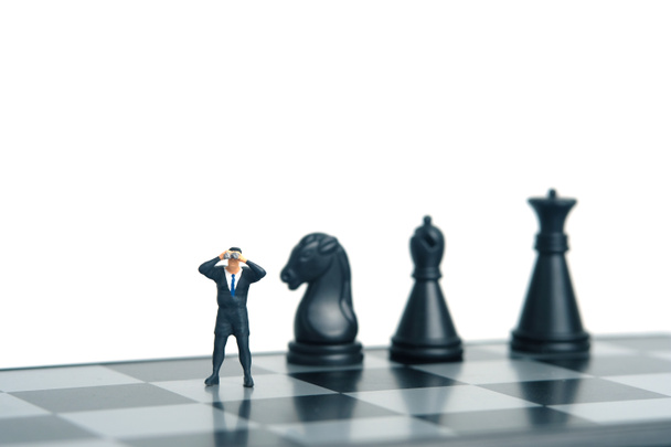 Επιχειρηματίας με κιάλια στέκεται δίπλα πιόνι σκάκι. Μικρογραφία μικροσκοπικοί άνθρωποι παιχνίδια φωτογραφία. απομονωμένο σε λευκό φόντο - Φωτογραφία, εικόνα