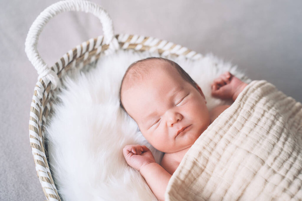 Sleeping newborn baby in basket wrapped in blanket in white fur background. Portrait of new born child one week old. - Φωτογραφία, εικόνα