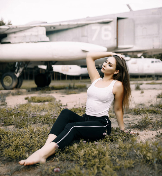 beautiful young brunette girl stands on the background of old military aircraft. Girl in a white shirt and black pants in nature. Military equipment. Portrait half-length. girl posing - Φωτογραφία, εικόνα