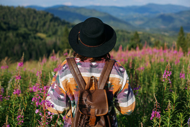 Žena cestovatel s batohem a kloboukem stojící v krásné divoké květiny a při pohledu na horské kopce. Wanderchtíč a pojetí cestování. Stylová ženská turistika v horách, atmosférický moment - Fotografie, Obrázek