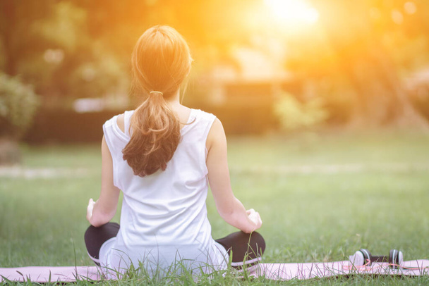Девушка сидит на коврике над газоном в парке, чтобы медитировать и просить благословения у Бога. Чтобы сохранить ее тело здоровым и медитации осанка является еще одной формой практики йоги, Верьте в Бога - Фото, изображение