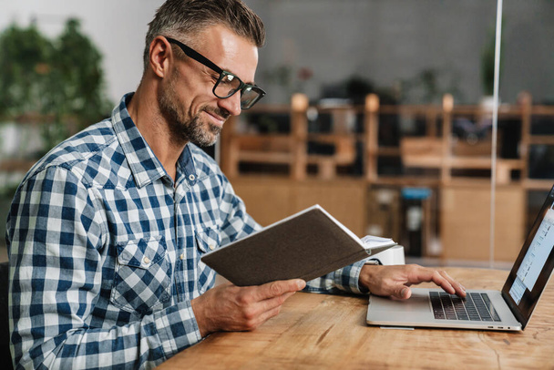 Boldog szürke férfi szemüveges olvasás közben dolgozik laptop az irodában - Fotó, kép