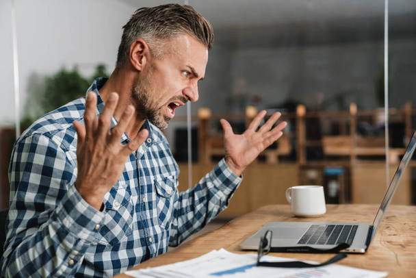 Furious grey man screaming and gesturing while working with laptop in office - Fotoğraf, Görsel
