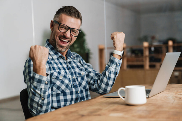 Excited man screaming and making winner gesture while working with laptop in office - 写真・画像