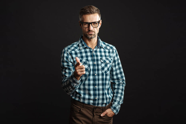 Handsome confident man in eyeglasses pointing finger and looking at camera isolated over black background - Foto, afbeelding