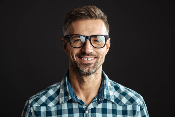 Handsome joyful man in eyeglasses smiling and looking at camera isolated over black background - Foto, afbeelding