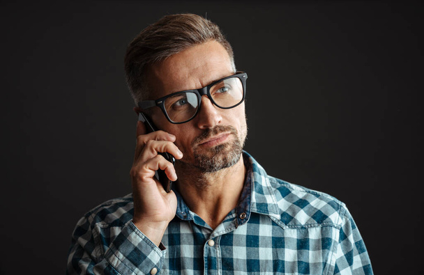 Photo of a concentrated serious grey-haired man talking by mobile phone isolated over grey wall background - Foto, Imagem