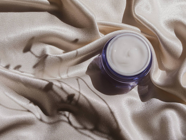 Blauw glas crème pot mockup op gladde zijde stof in zonlicht met bloemen schaduw Natuurlijke huidverzorging moisturizer cosmetische, gezonde gloeiende huid SPA wellness gezicht lichaam hydratatie behandeling UV-bescherming - Foto, afbeelding