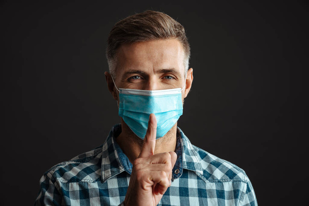 Photo of pleased grey-haired man wearing face mask isolated over grey wall background and showing silence gesture - Zdjęcie, obraz