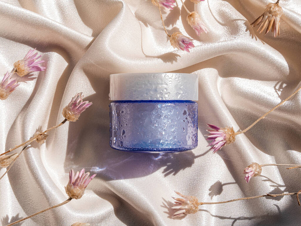 Blue glass cream jar mockup on smooth silk fabric sunlight flowers splashes water. Увлажняющий увлажняющий крем для кожи косметический, здоровый SPA оздоровительный увлажняющий уход за кожей лица УФ-защиты - Фото, изображение
