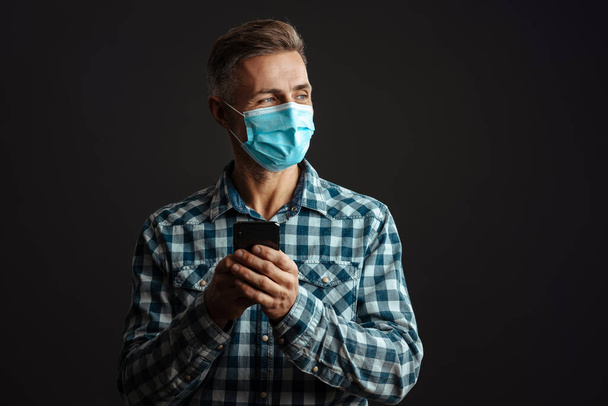 Photo of optimistic grey-haired man wearing face mask and using mobile phone isolated over grey wall background - Zdjęcie, obraz