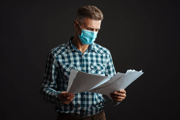 Photo of serious grey-haired man wearing face mask isolated over grey wall background and holding documents - Foto, Imagen