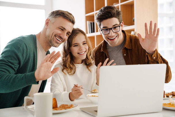 Happy gay couple and adopted child on a video call via laptop computer at home, waving hands during breakfast - Фото, зображення