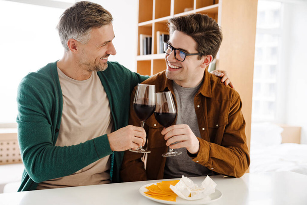 Happy caucasian gay couple hugging while drinking wine at home - Foto, imagen