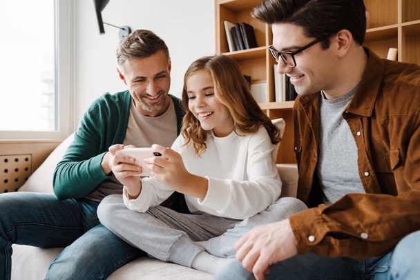 Happy gay family smiling and using mobile phone while sitting on couch at home - Φωτογραφία, εικόνα