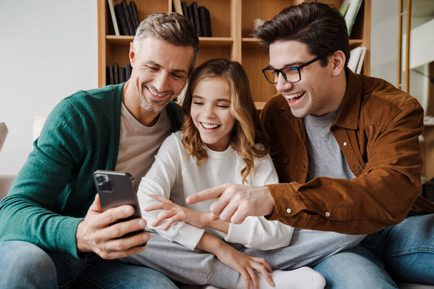 Happy gay family smiling and using mobile phone while sitting on couch at home - Fotografie, Obrázek