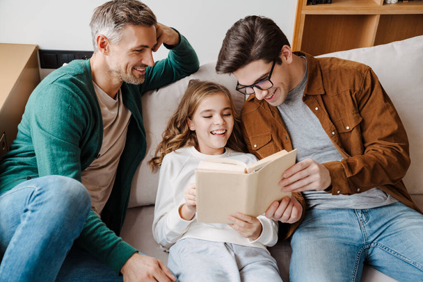 Happy gay parents reading book together with their little daughter at home - Foto, Bild