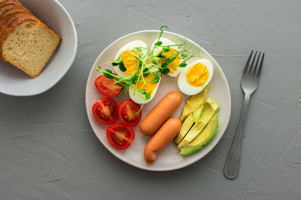 Вкусный завтрак, вареные яйца с сосисками, помидоры и авокадо, микрозелень гороха и подсолнухов, тарелка с едой и миска хлеба на сером фоне, вид сверху - Фото, изображение