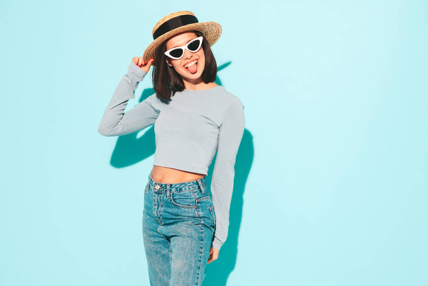 Portrait of young beautiful smiling female in trendy summer hipster  clothes. Sexy carefree woman posing near blue wall in studio. Positive model having fun in sunglasses and hat. Cheerful and happy - Photo, Image
