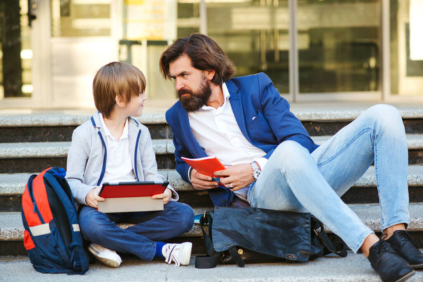 Ο μπαμπάς και ο γιος κάθονται στις σκάλες πριν το σχολείο. Χαριτωμένο αγόρι και μοντέρνος μπαμπάς διασκεδάζουν μαζί. Ο πατέρας μιλάει με τον γιο του μετά τα μαθήματα εκτός σχολείου. Σχολιαρόπαιδο με σακίδιο και σημειωματάρια. - Φωτογραφία, εικόνα