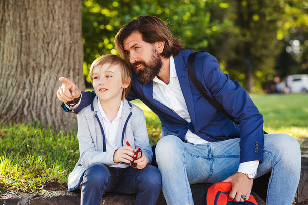 Cute boy talking with dad on a walk after school. Bearded man and his son outside. Happy father and son spending time together. Back to school concept. Family, relationship and lifestyle concept. - Фото, изображение