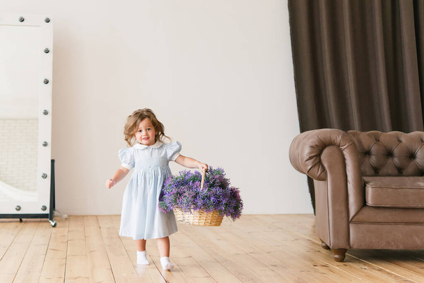 Little girl with a basket of flowers. Dressed in a simple blue dress. In room. - Foto, Imagem