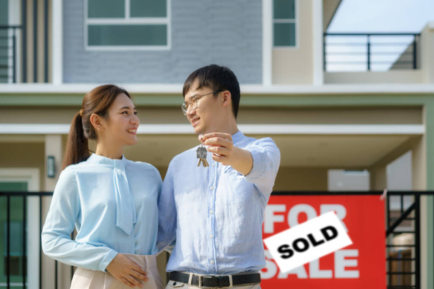 Portrait of Asian young couple standing and hugging together and holding house key looking happy in front of their new house to start new life. Family, age, home, real estate and people concept - Photo, Image
