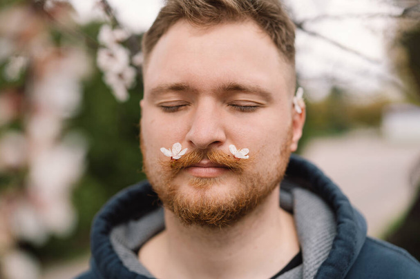 Άντρας με άσπρα λουλούδια στο μουστάκι. Ανέπνευσε. Ευτυχισμένος άνθρωπος χωρίς αλλεργία. Ανοιξιάτικη αλλεργία. Άνοιξη - Φωτογραφία, εικόνα