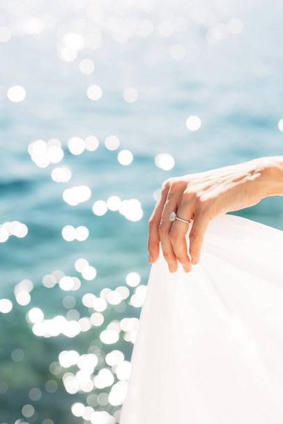 Die Hand der Braut mit einem Ehering am Finger, in Großaufnahme. Die Braut hält den Rand des Kleides vor dem Hintergrund des Meeres.  - Foto, Bild