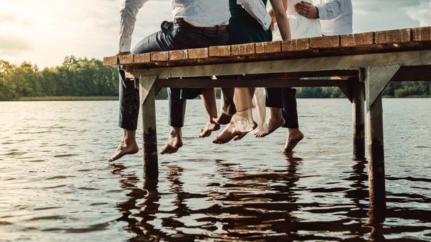 Un grupo de amigos sentados en el muelle de madera con los pies en el lago - Foto, imagen
