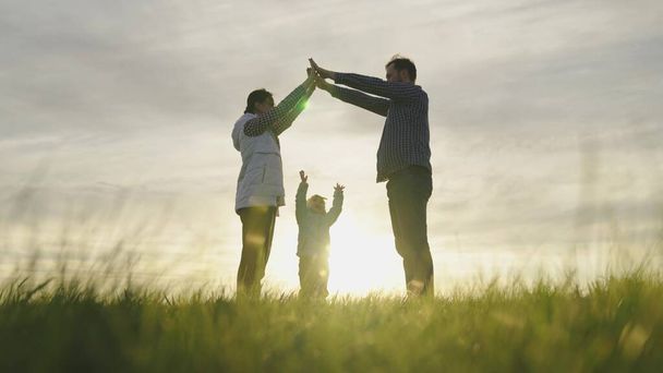Onnellinen perhe leikkii pienen tyttären kanssa, perheen symboli kotona auringossa. Äiti on isä ja he kuvaavat käsillään talon katon lapsen pään päälle. Unelma yhteisestä kodista. Kodinrakentajat - Valokuva, kuva
